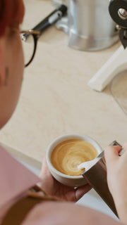 Vertical shot of female barista in uniform adding milk from pitcher to cup while preparing coffee for serving in coffee shop
