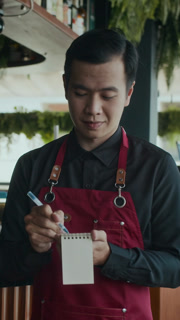 Vertical shot of waiter of Asian ethnicity in apron making notes while listening to client ordering in cafe