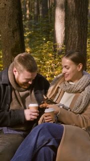 Vertical shot of affectionate young couple kissing, drinking coffee and petting cute puppy while sitting on bench during walk in fall forest park