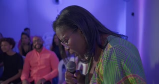 Woman speaks into microphone with bowed head in emotional moment during church service, congregation seated and watching with reverence
