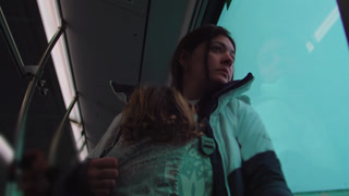 Mother standing by bus window holding sleeping toddler in front carrier during rainy commute showing protective care bonding and urban travel routine