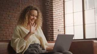 Woman sits on sofa, waves hello on video call