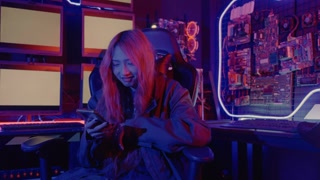 Portrait of happy young Asian woman using mobile phone while sitting in front of a computer monitor display in neon light.