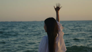 Back view of Asian woman on beach waving hand in goodbye gesture to ocean, looking at calm sea water with arm raised