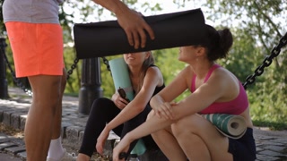 After finishing a yoga session, three friends sit on the in the park, smiling and chatting. One is holding a yoga mat, another is scrolling
