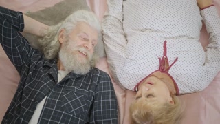 Top view senior couple lying in bed in pajamas talking and smiling. Happy carefree loving Caucasian man and woman enjoying weekend morning at home indoors. Slow motion.