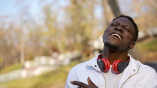 Confident joyful African American man singing standing in sunny autumn park. Portrait of happy carefree guy enjoying leisure on weekend outdoors. Live camera panning.