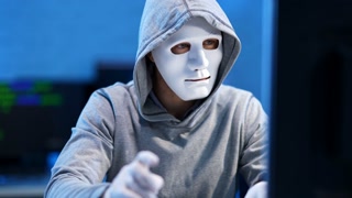 Caucasian hacker in white mask and gloves coding indoors and waving at camera. Unrecognizable man hacking internet website or coding computer virus. Programming concept.