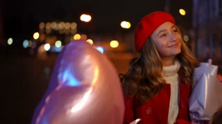 Satisfied Caucasian teenage girl walking in night city with bouquet of roses and heart-shaped balloon. Portrait of happy relaxed teenager strolling in city looking around on Valentine's.