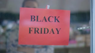 Blurred woman behind glass placing Black Friday announcement on showcase. Unrecognizable young Caucasian seller advertising sales in shoe shop indoors.