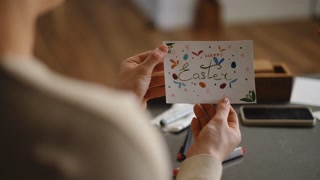 Slow motion. A woman holds an Easter greeting card that she drew with colored markers and watercolors while sitting at a table in the living room. DIY Easter card. Over-the-shoulder shot.