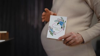 Close-up. Slow motion. Happy Mother's Day. Profile of an unrecognizable pregnant woman, holding a Mother's Day card with one hand and rubbing her belly with the other hand, while standing indoors.