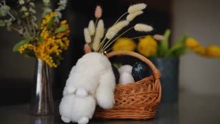 Close-up. Celebration of Easter. On the table in the apartment there is a glass vase with spring flowers, an Easter bunny, and a basket with Easter eggs. A woman's hand places a handmade Easter card near a basket.