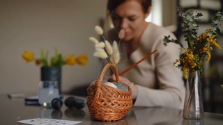 Slow motion. Close-up. A woman carefully ornaments watercolor on the Easter egg with a paintbrush while sitting at a table in the living room. Preparations for the celebration of Easter.