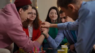 Young Caucasian men wrestling arms as joyful friends cheering supporting mates. Happy relaxed confident guys having fun on party indoors.