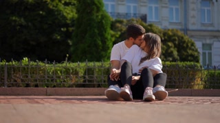 Wide shot portrait of loving happy relaxed couple kissing sitting on the right on skateboard outdoors. Relaxed confident Caucasian boyfriend and girlfriend dating on city street hugging cuddling.