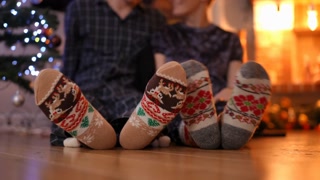 Close-up male and female feet in socks with Christmas pattern shaking as blurred man and woman talking sitting at New year tree at background. relaxed happy Caucasian couple on holiday at home.