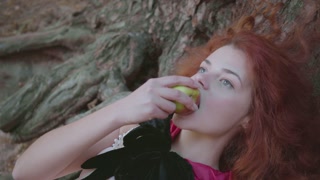 Red-haired woman in black gown enjoying apple under tree. Witch relaxing in autumn forest. Snow White vibes.