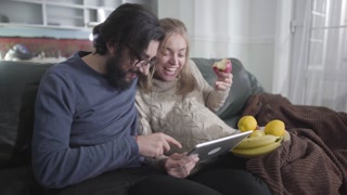 Cheerful married couple looking at tablet screen and laughing out loud. Pregnant Caucasian woman showing her husband something funny and eating apple. Family enjoying time together indoors.