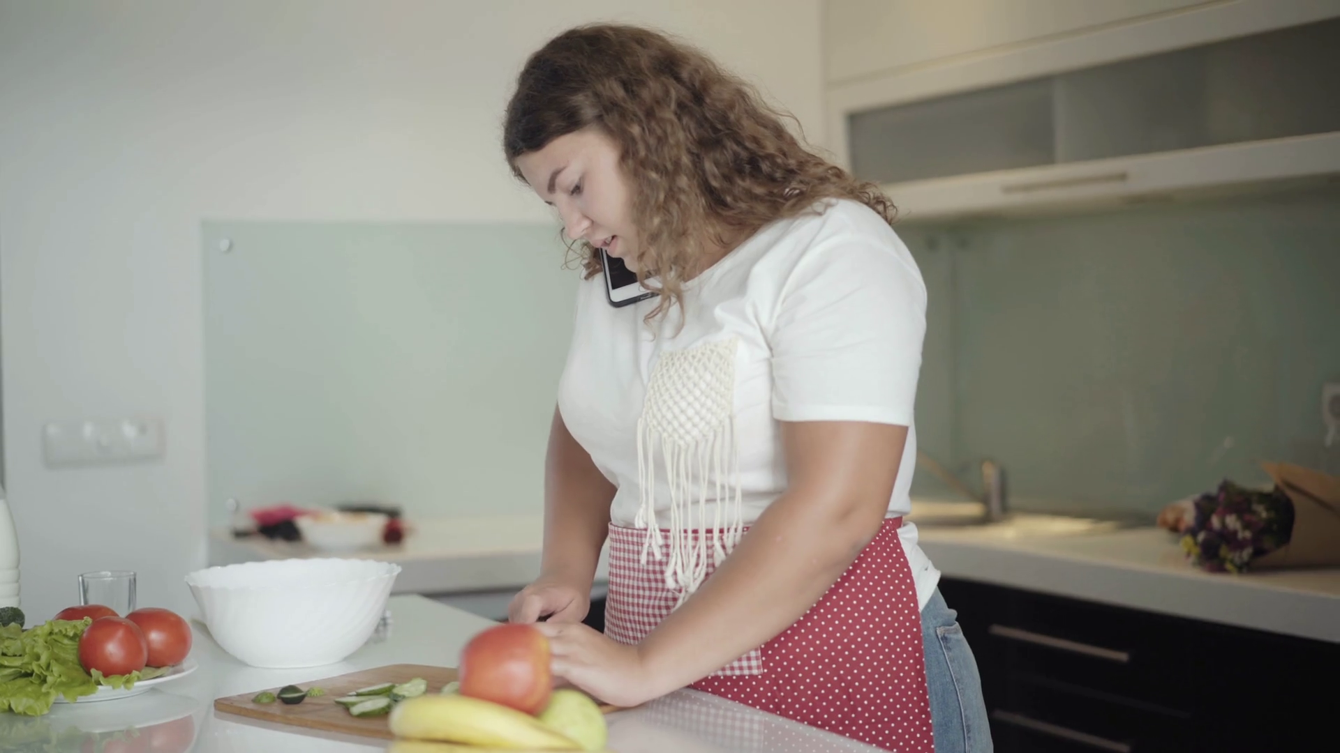 Chubby woman talking on the phone in kitchen as cooking fresh healthful salad. Portrait of positive young plus-size Caucasian lady at home in the morning image