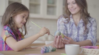 Daughter painting Easter egg with brush, woman smiling. Mother-daughter bond during Easter prep.