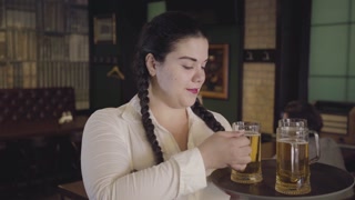Plump waitress with pigtails in white blouse sneaks a drink from a customer's glass. Funny girl can not resist and drink delicious beer. Leisure at the bar