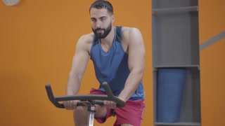 Portrait of concentrated muscular Middle Eastern man riding exercise bike in gym. Confident strong sportsman exercising in sports club. Fitness, sport, masculinity, healthy lifestyle.