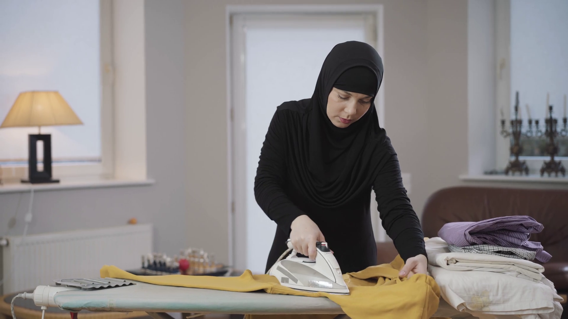 Portrait of concentrated Muslim woman in hijab ironing yellow pullover