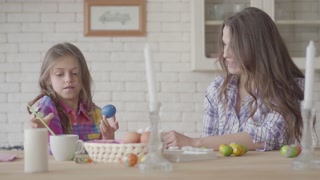 Mother and daughter at the Easter table. Girl paints an Easter egg. Family bonding during Easter preparations.