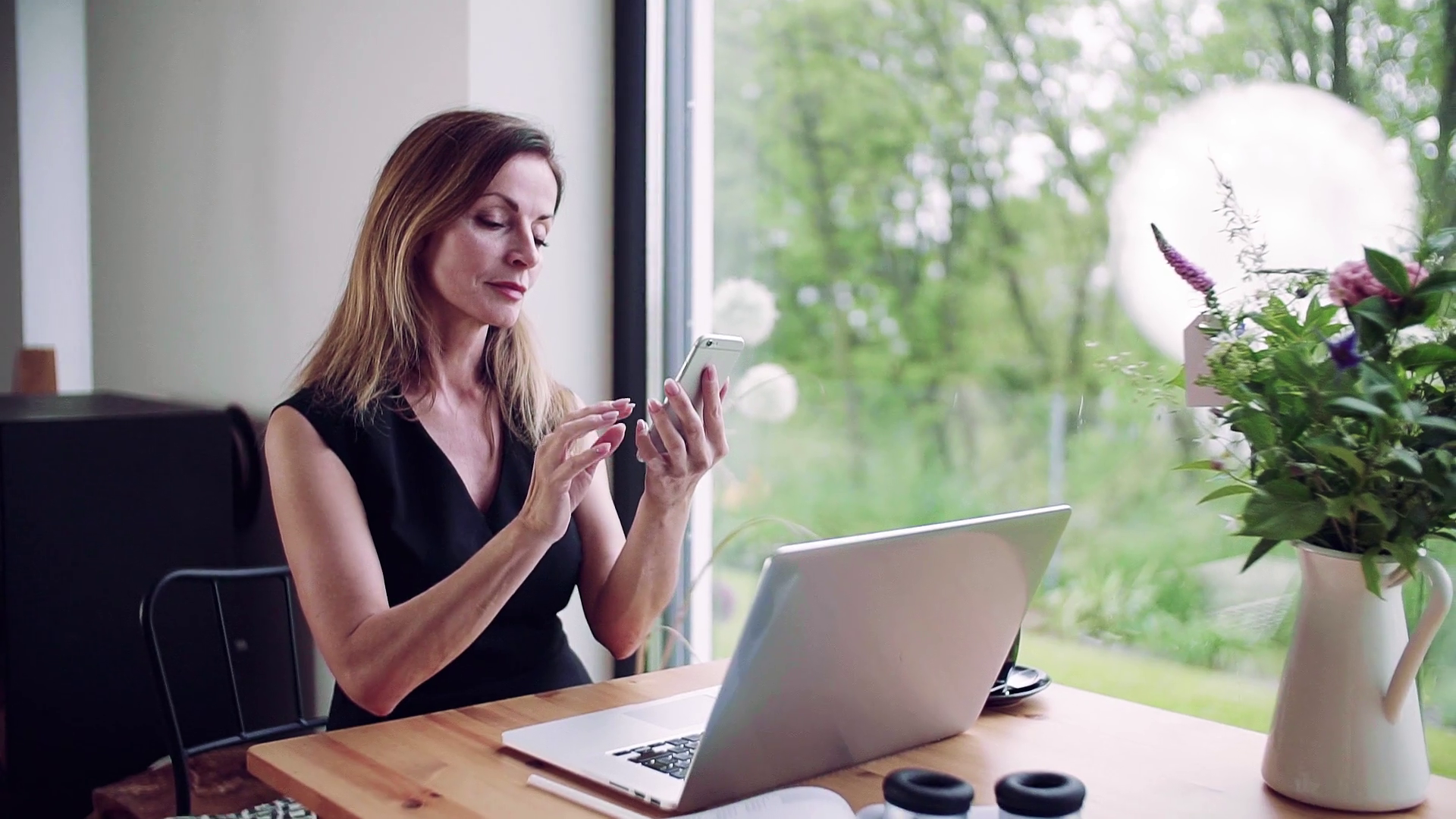 A mature woman with laptop sitting at the table, using smartphone in home office pic