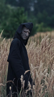 Scary black figure on a wheat field. Terrible ghost in dark mantle with hood looking at camera and turning around on nature evening background. Horror concept. Vertical video