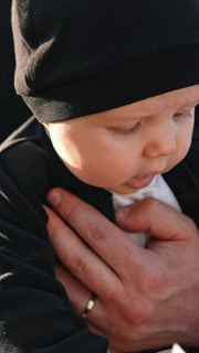 Cute little infant wearing black suit and cap. Baby in hands of parents is interested in the box in front of him. Close up. Vertical video