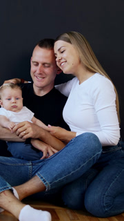 Young modern family wearing jeans sit on the floor with baby in hands. Happy couple kissing. Black backdrop. Vertical video