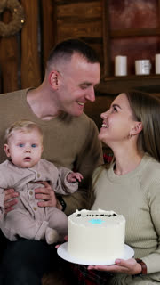 Loving Caucasian couple kissing each other tenderly. Woman holds a cake in hands and man holds a cute baby. Christmas trees at backdrop. Vertical video