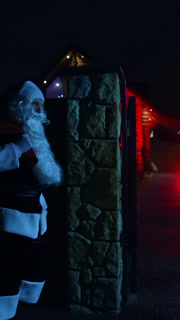Santa Claus with red bag at night. Santa walks into front yard of house decorated with lights and garlands. Santa carrying gifts for children. Vertical video