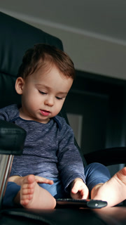Little baby boy playing with tv remote sitting in the black office chair. Calm child with bare feet playing at home. Low angle view. Vertical video