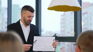Bearded businessman wearing black suit holds a chart in his hands. Boss listens to his employees' ideas about the graphs.