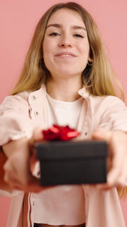 Smiling young woman offering a gift wrapped in black paper with a red bow, isolated on a pink background