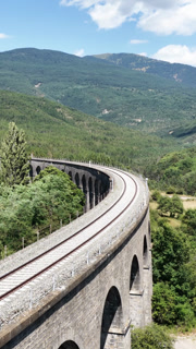 Empty railway tracks running along a historic stone arch viaduct curving through a lush green mountain landscape on a sunny day with blue