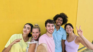 Happy multicultural group of friends taking a selfie video against a yellow wall. Young people laughing, smiling, and making funny faces