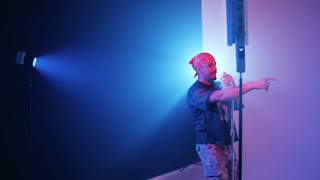 Charismatic male rapper performing a reggaeton song in a studio. The artist is singing passionately into a microphone under blue stage
