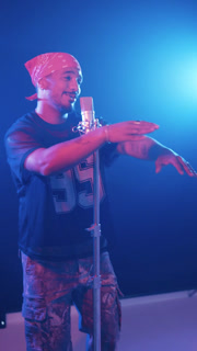 Male hip hop artist performing a song in a recording studio with blue and pink neon lights. Another person is filming with a smartphone