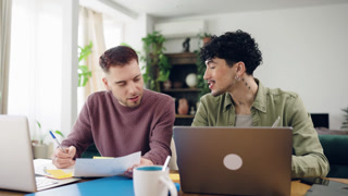 Young gay couple feeling overwhelmed while working on their finances at home. One man is using a laptop, feeling stressed, while his partner