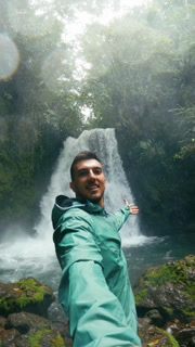 Young male content creator filming himself with a beautiful waterfall in the background at Arenal Volcano National Park, Costa Rica