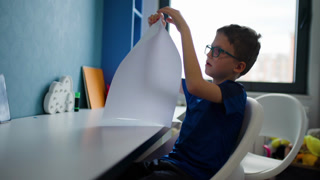 Boy Folds Paper at Desk: A Child with Glasses Engrossed in a Quiet Activity in His Bedroom