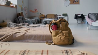 No people medium closeup of backpack with thermos cup next to mattress with blanket on floor in refugee shelter. Folding beds, bags with personal possessions, information board in background
