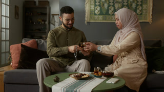 Front view shot of young Muslim couple enjoying teatime together in cozy modern home with woman wearing traditional attire pouring tea for husband
