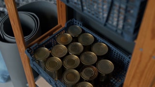 High-angle medium no people shot of box with canned foods on shelf in emergency refugee accommodation facility, next to rolled-up camping mats and blankets