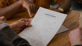 Medium closeup of hands of unrecognizable mature friends sitting at table in cafe and choosing food for lunch. Woman wearing chunky silver rings pointing at menu and discussing meals with companions
