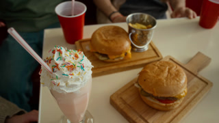 High-angle close-up of sweet milkshake with whipped cream and straw, cheeseburgers, potato fries in bucket and glasses of soda served on table for young people socializing at fast food restaurant